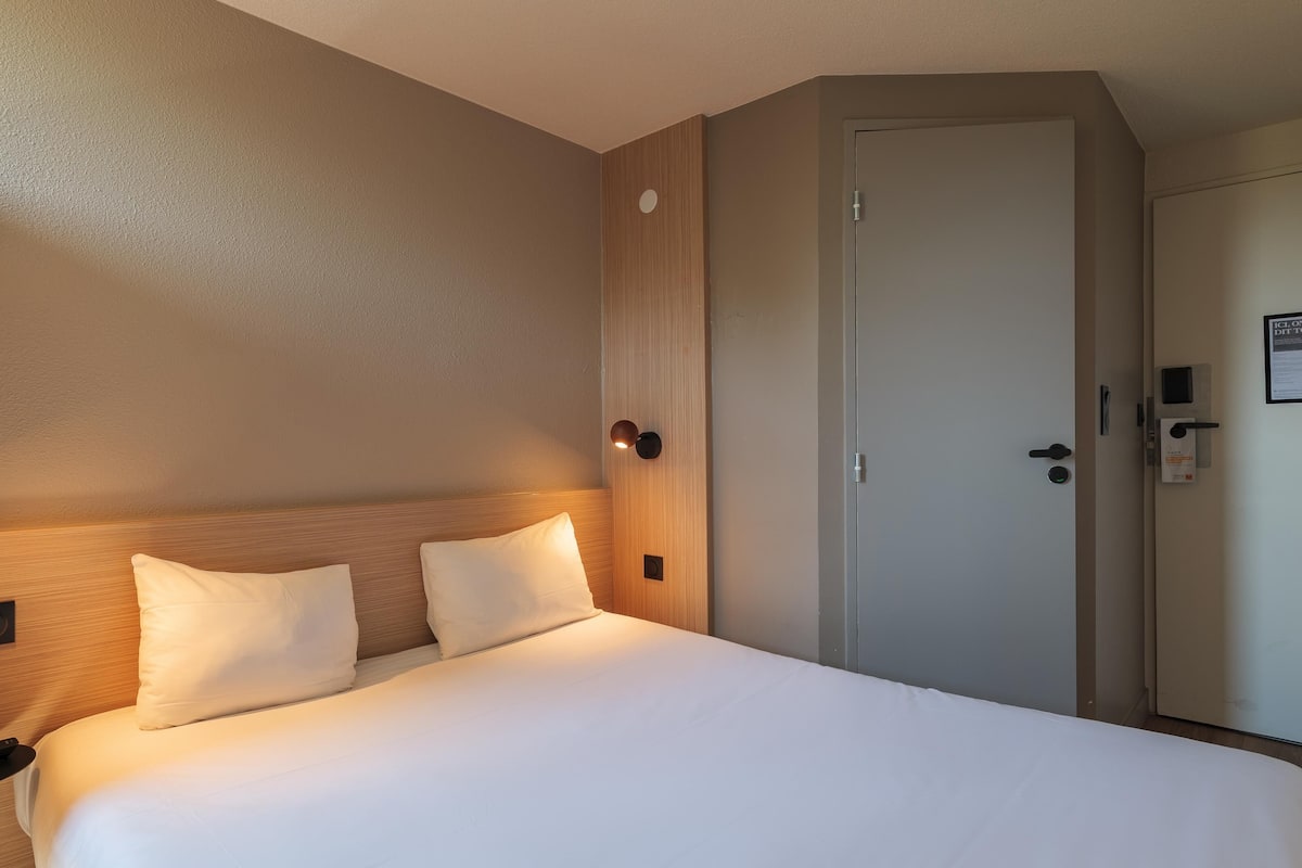 Quarto de hotel com uma cama, duas luminárias de cabeceira, uma porta e um painel de controle fixado na parede.
