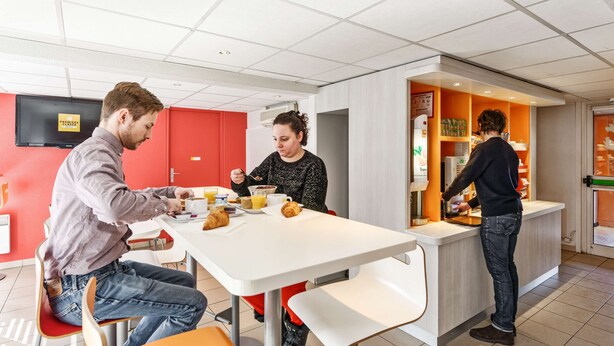 Ein moderner Essbereich mit einem weißen Tisch, roten Wänden und einer Person, die an einer Arbeitsplatte etwas zubereitet.