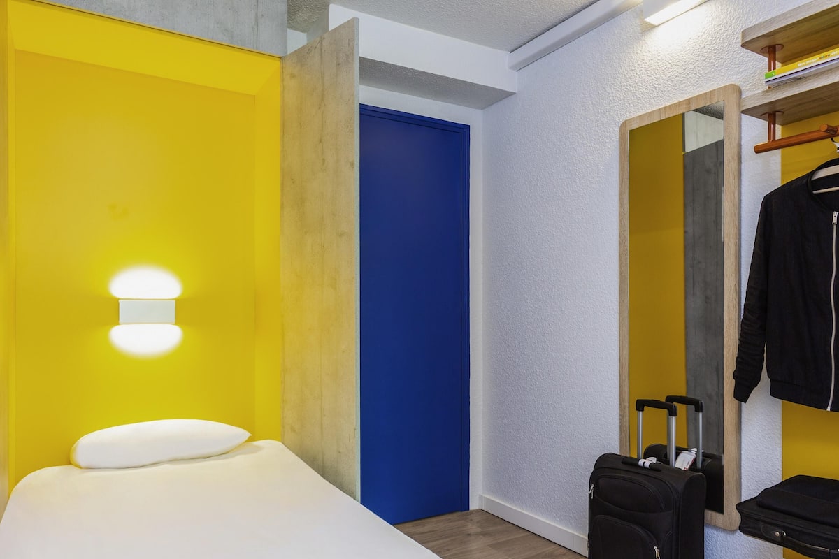 A hotel room with a bed, a blue door, a mirror, and a suitcase.