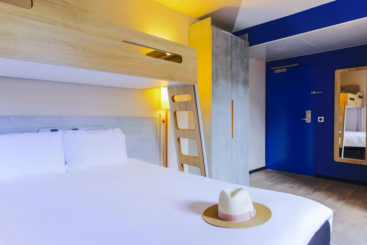 A hotel room with a bunk bed, a ladder, a white bedspread, a hat on the bed, and a blue door.