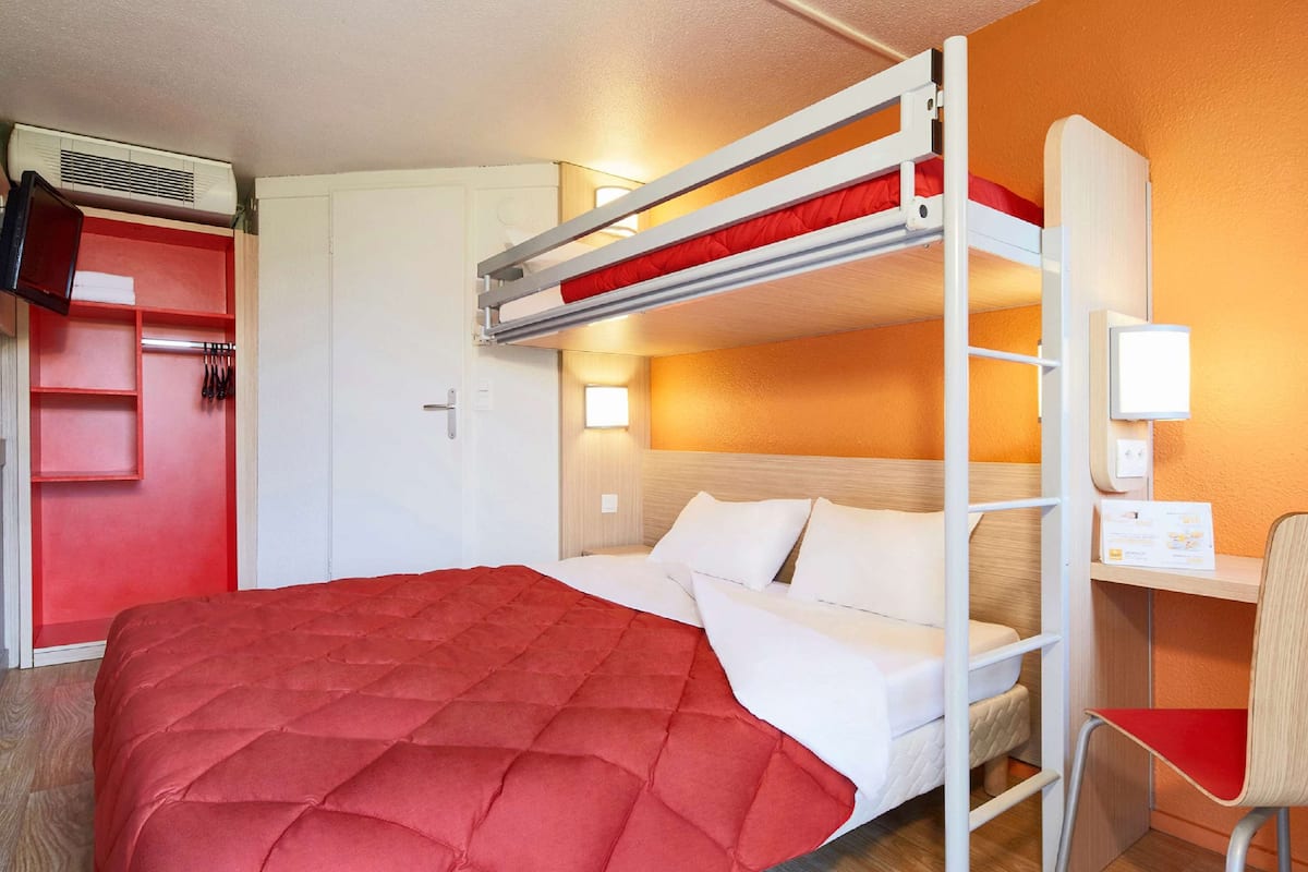 A bunk bed with a red quilt, a wooden desk, a chair, and a wardrobe with a red door.