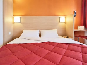 Une chambre d’hôtel avec une tête de lit en bois, deux lampes de chevet, une couvre-lit rouge matelassée et une petite table avec une télécommande.