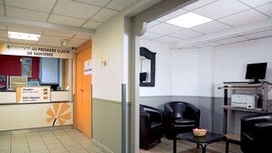 Zona de recepción con un cartel que dice “Bienvenue au Premiere Classe de Nanterre” y un mostrador con una computadora e impresora. Hay sillas de cuero negro y una pequeña mesa con un frutero.