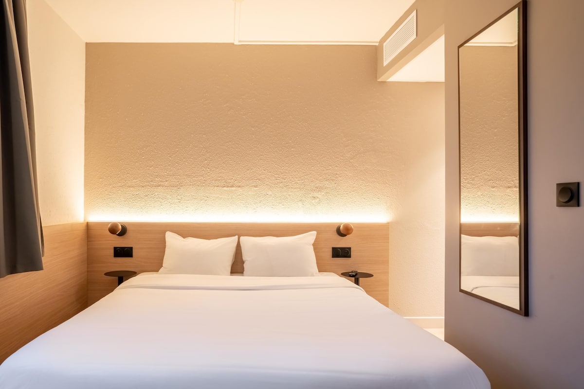 Una habitación de hotel con una cama grande, un cabecero con iluminación integrada, un espejo y una ventana con cortinas.