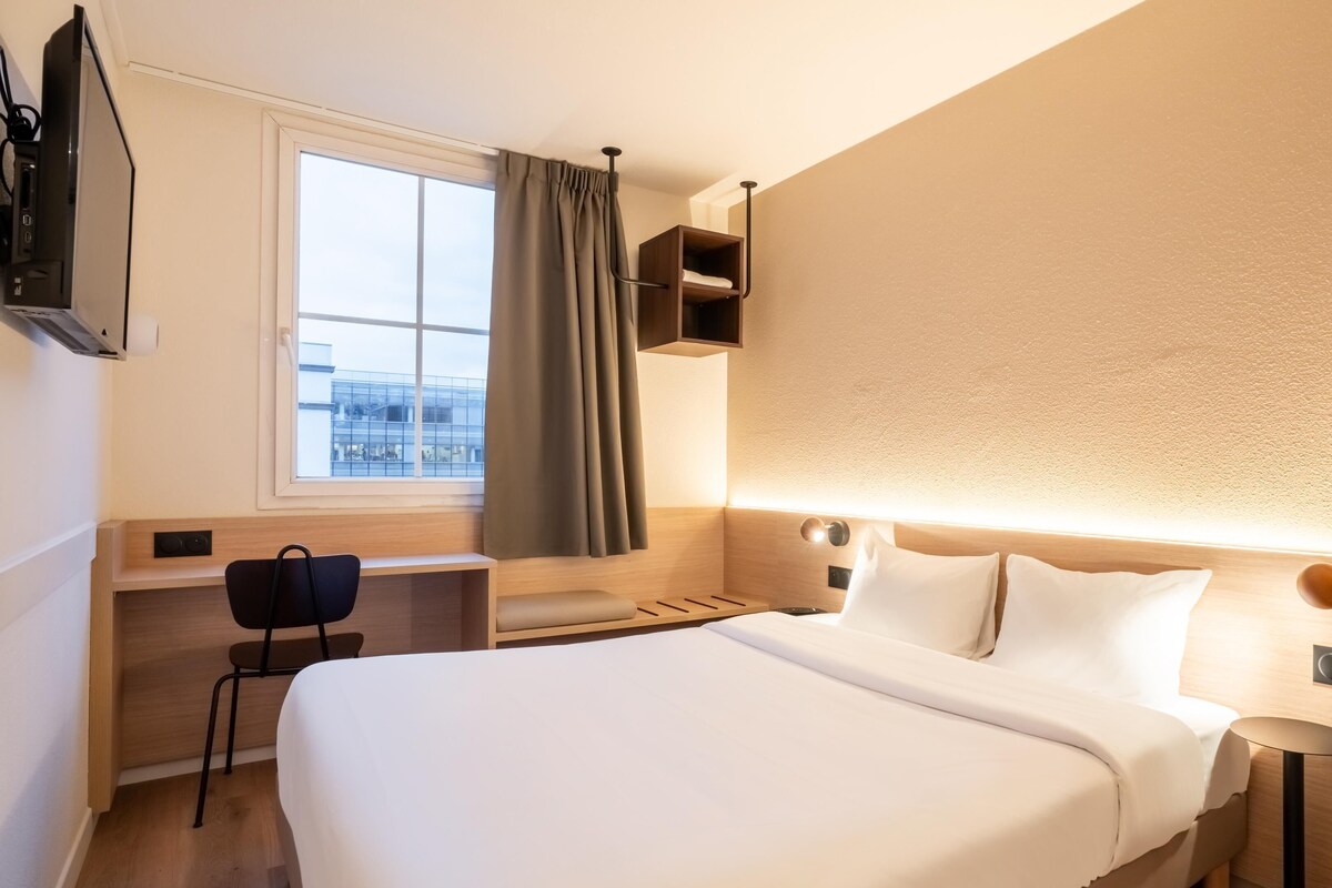 A hotel room with a large bed, a desk with a chair, a window with curtains, and a wall-mounted shelf.