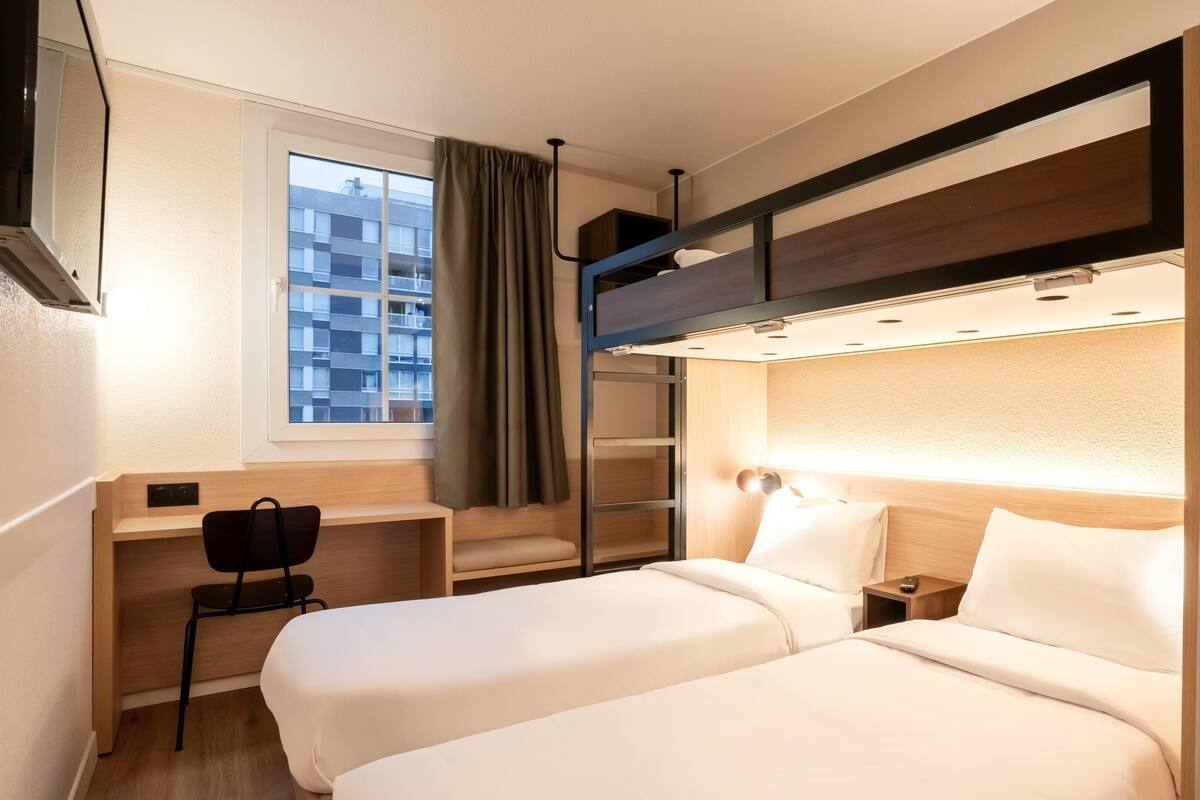 Habitación de hotel con dos camas, un escritorio, una silla, un ventanal con cortinas y una litera.