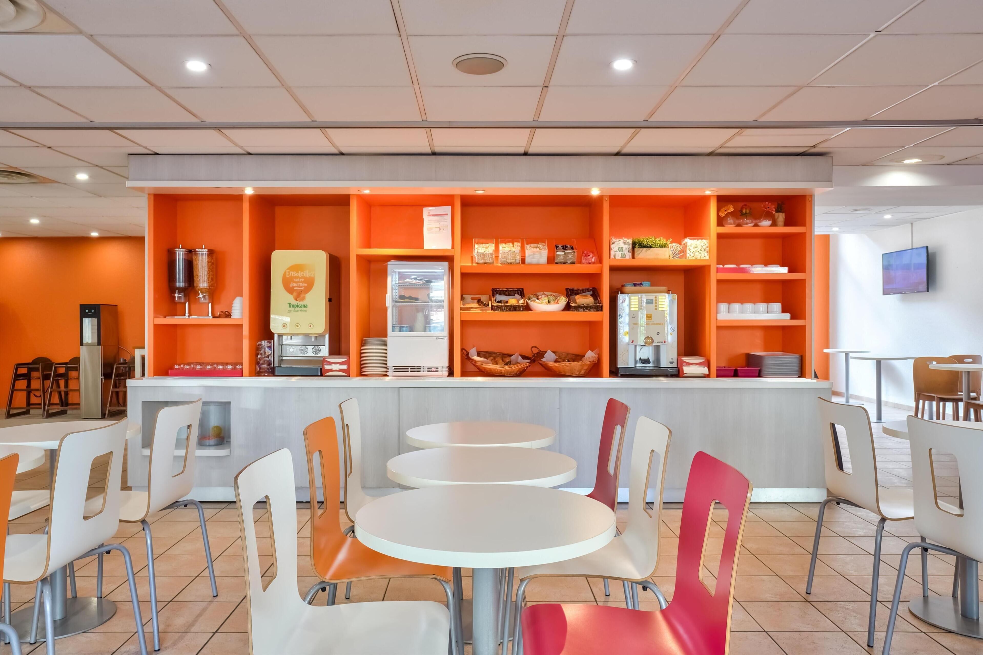 Un café moderno con decoración en naranja y blanco, que incluye mesas y sillas redondas, una máquina de café y diversos alimentos en exhibición.