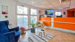 Zona de recepción de un hotel con una silla azul, un mostrador de recepción, un televisor y una planta en maceta.