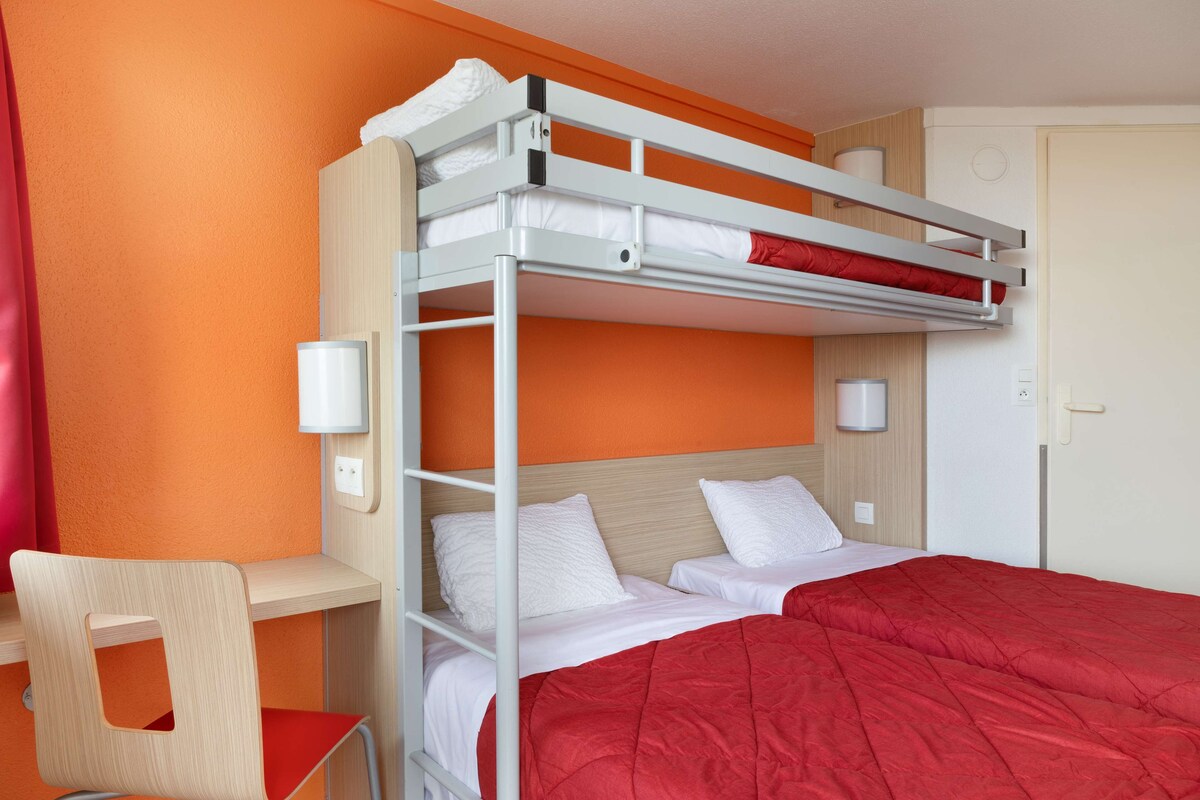 Una cama litera con ropa de cama blanca y una manta roja, un escritorio de madera y una silla con cojín rojo en el asiento.