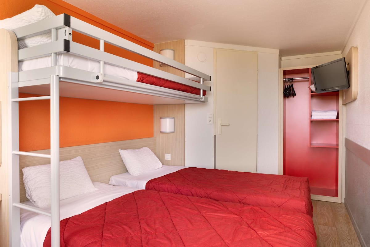 A bunk bed room with a red bedspread, a television mounted on the wall, and a wardrobe with hanging clothes.