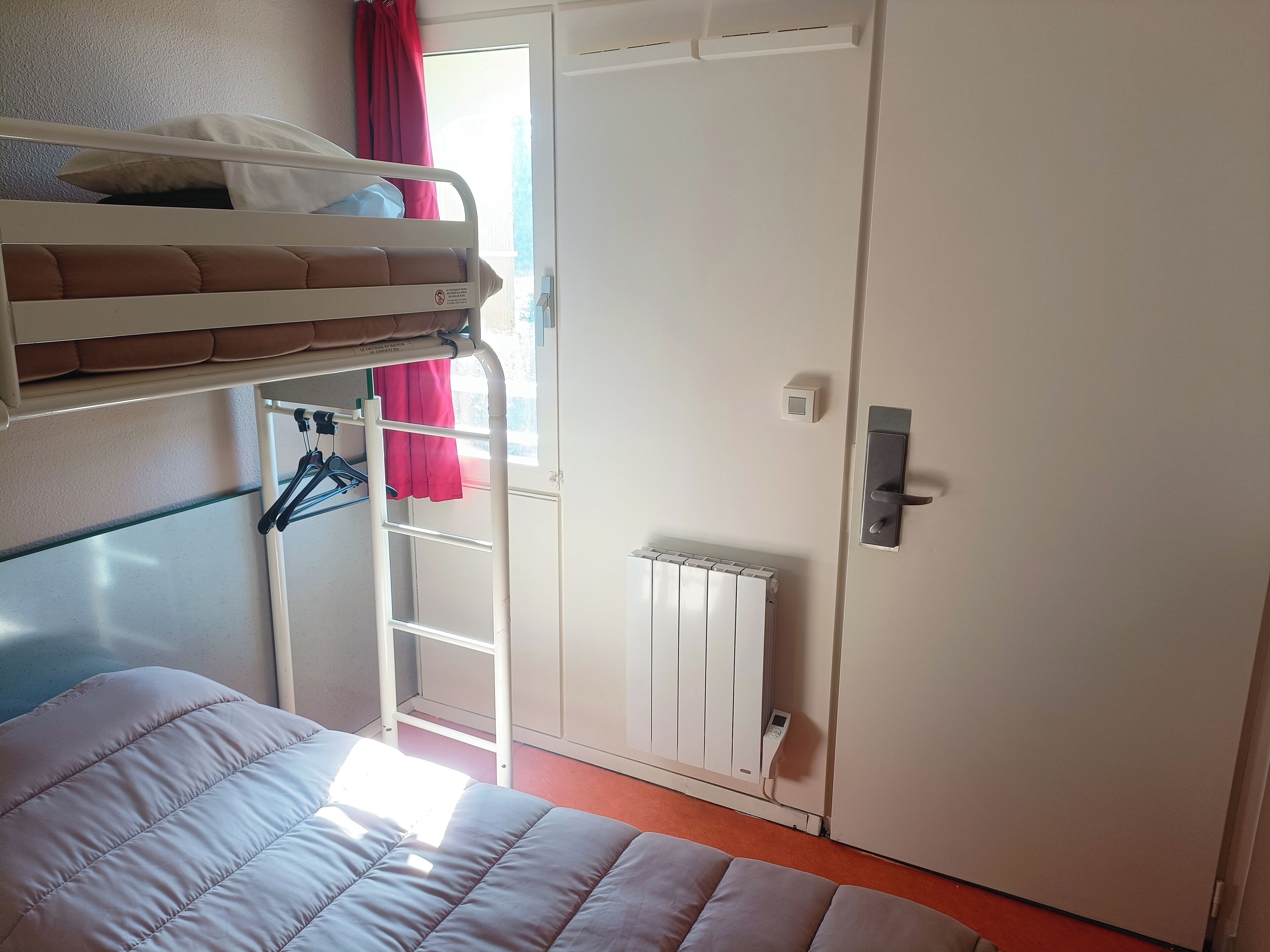 A bunk bed with a white headboard, a radiator, a door with a handle, and a window with a red curtain.