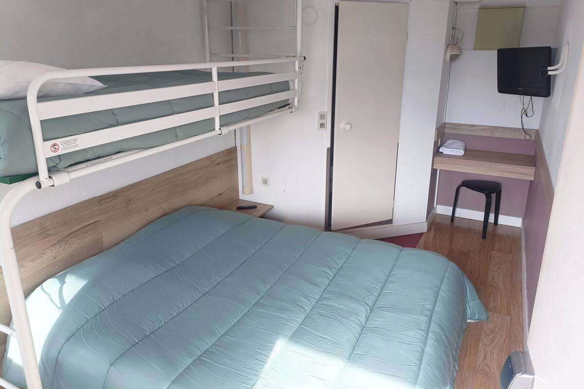 Une chambre avec un lit superposé, un bureau et une chaise, une porte et un petit tabouret.