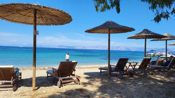 A beach with wooden lounge chairs and thatched umbrellas, a clear blue sea, and distant mountains. - Helios Splendid (Corfu)