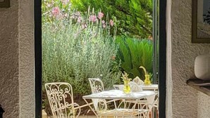 An arched doorway leading to an outdoor seating area with white metal chairs and a table, surrounded by lush greenery and flowering plants. - Helios Splendid (Corfu)