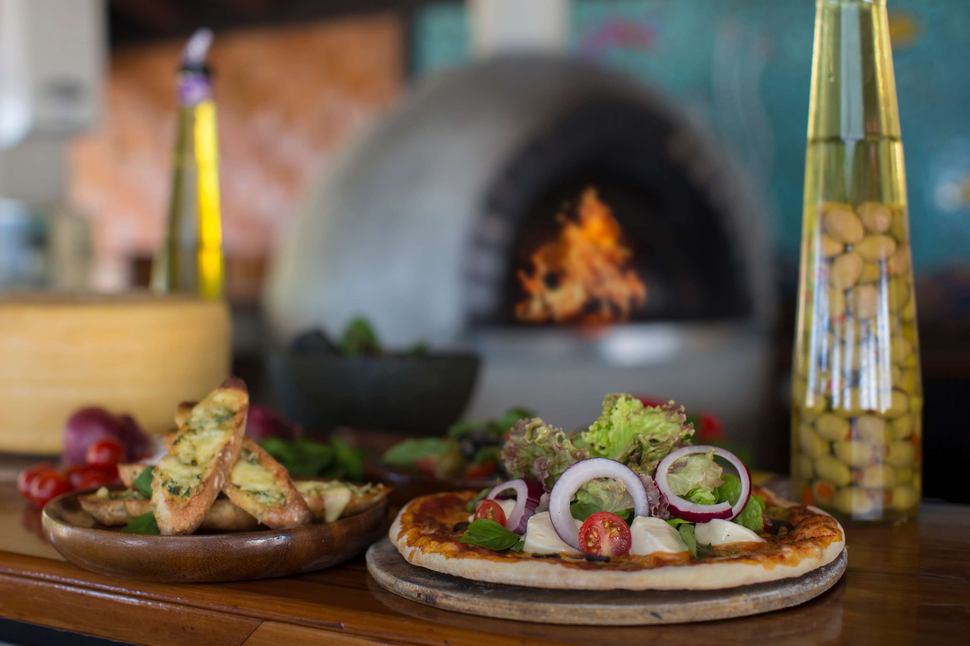 Two pizzas on a wooden table with a pizza oven in the background.