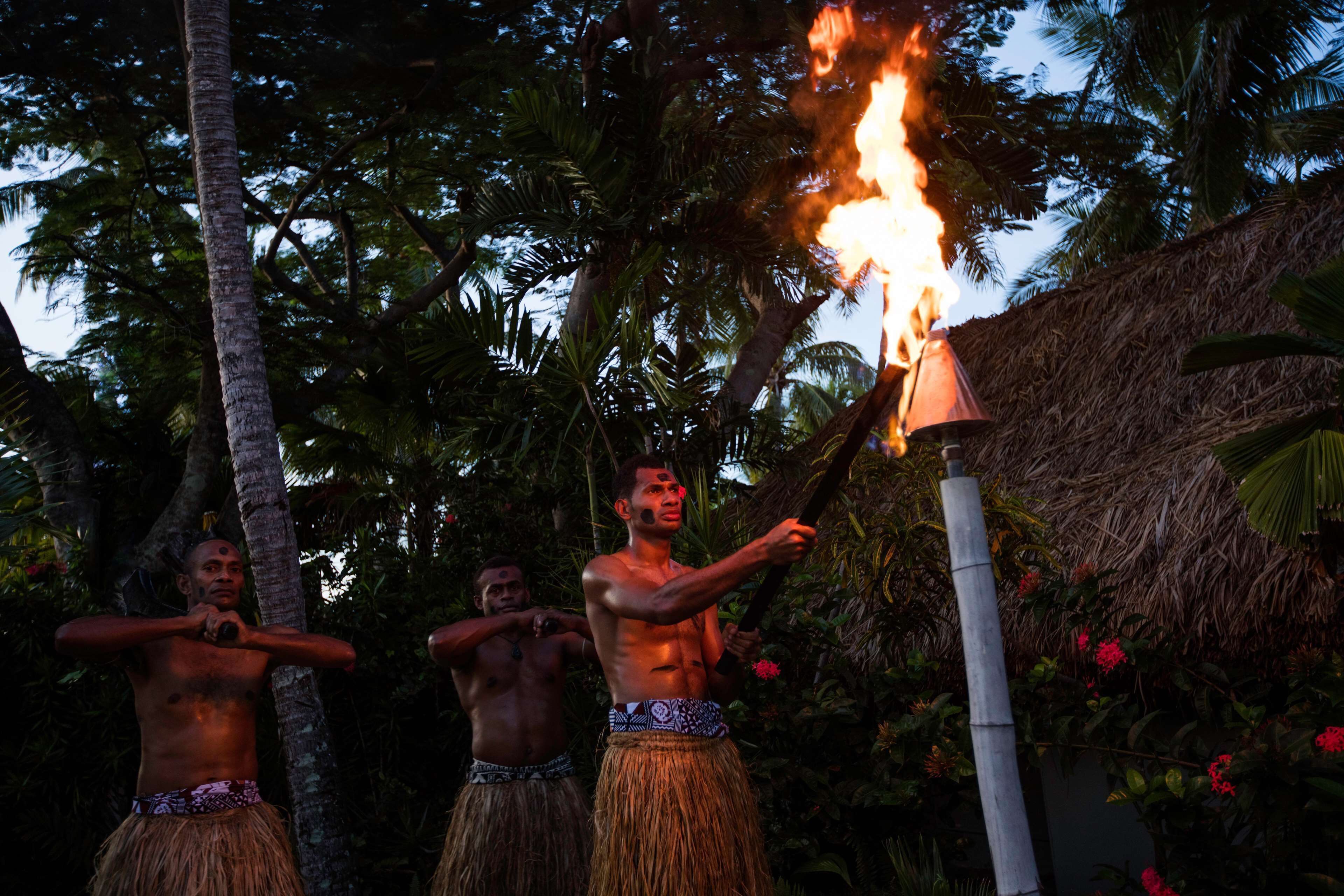 Three individuals performing a fire ritual with a torch in a tropical setting.