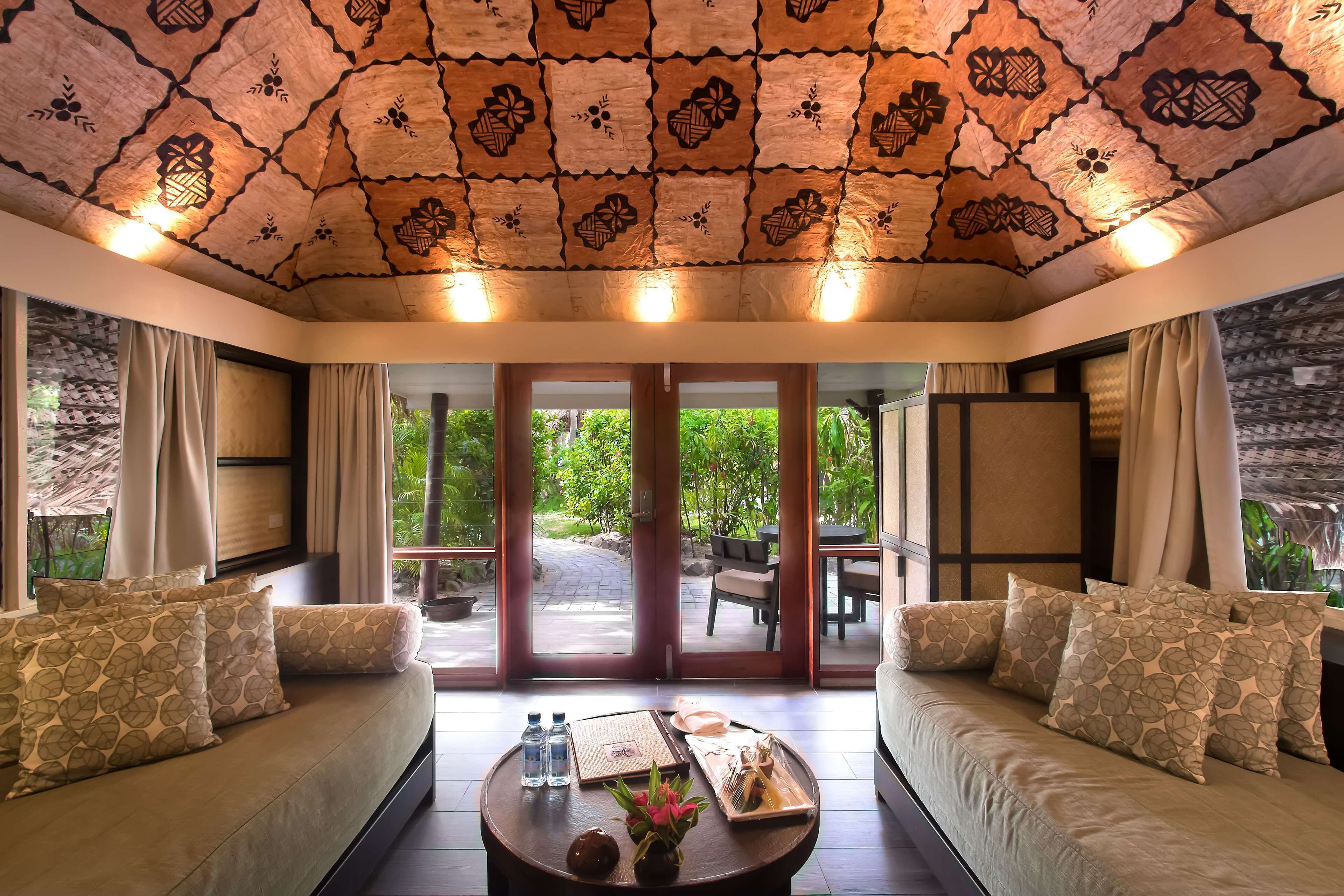 A room with a patterned ceiling, a sofa with patterned cushions, a coffee table with a tray of food and drinks, and a view of greenery through the glass doors.