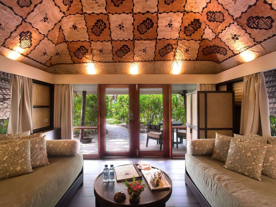 A room with a patterned ceiling, a sofa with patterned cushions, a coffee table with a tray of food and drinks, and a view of greenery through the glass doors.