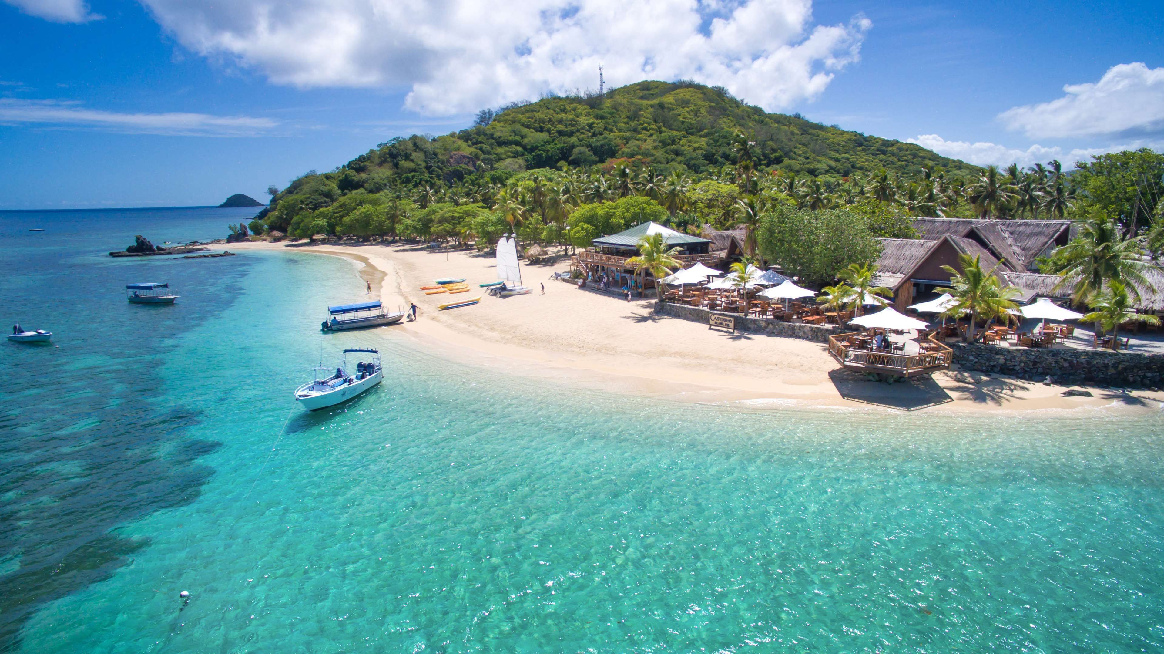 Un resort en una playa tropical con aguas turquesas cristalinas, barcos y una exuberante colina verde al fondo.