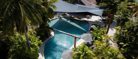 A swimming pool surrounded by palm trees and a thatched-roof building.