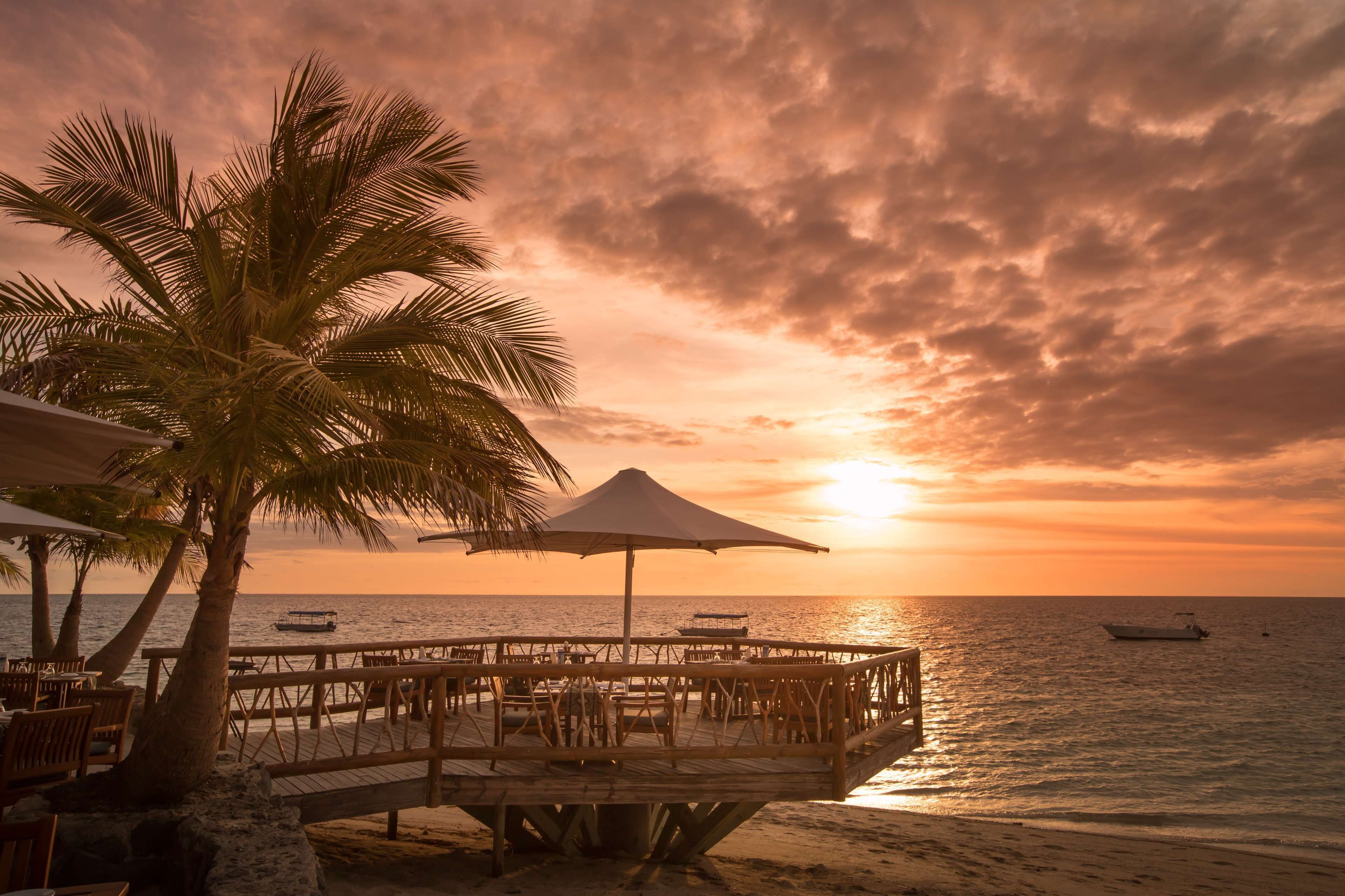 A wooden deck with tables and chairs, overlooking the ocean at sunset.