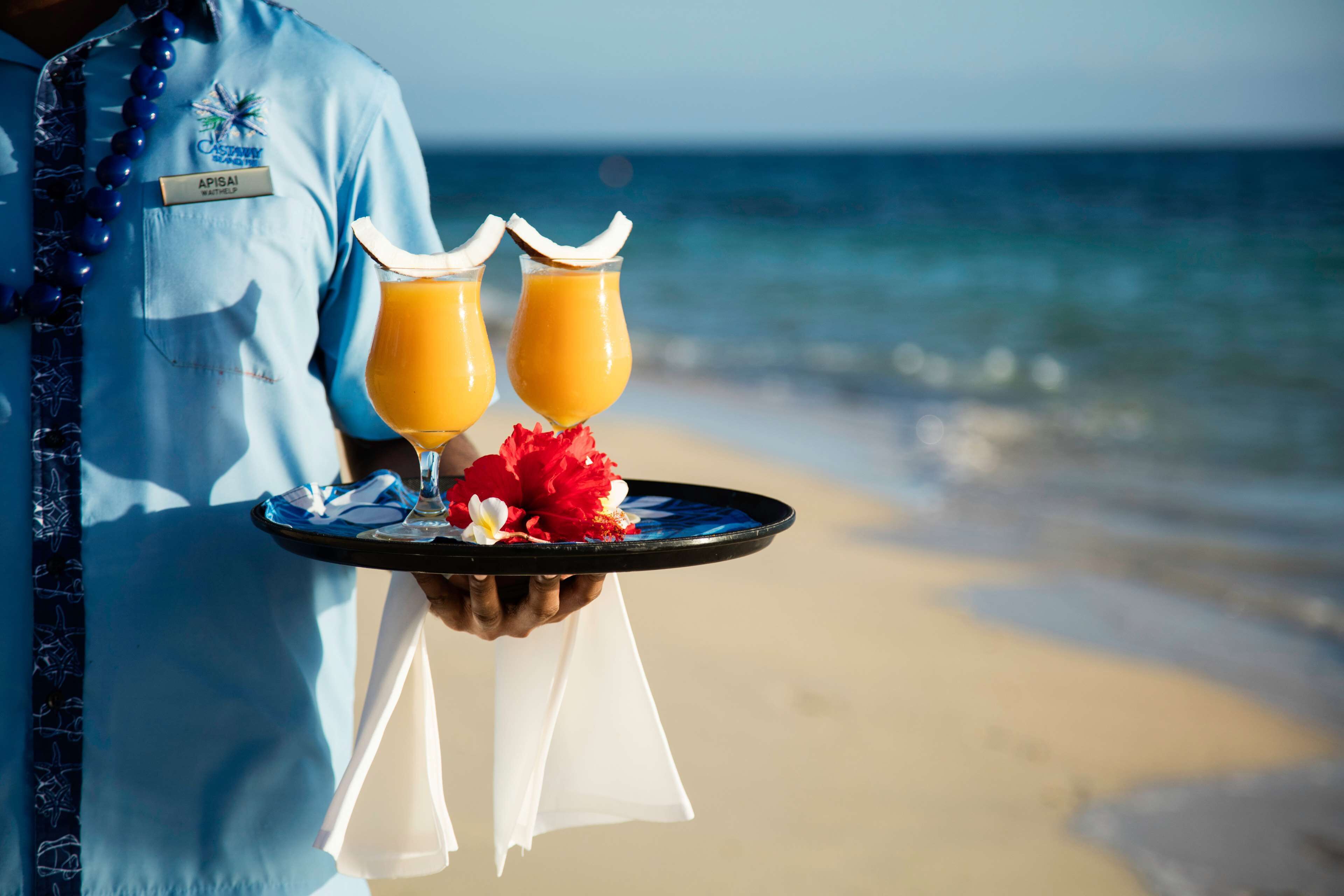 A person holding a tray with two drinks and a flower, standing on a beach with the ocean in the background.