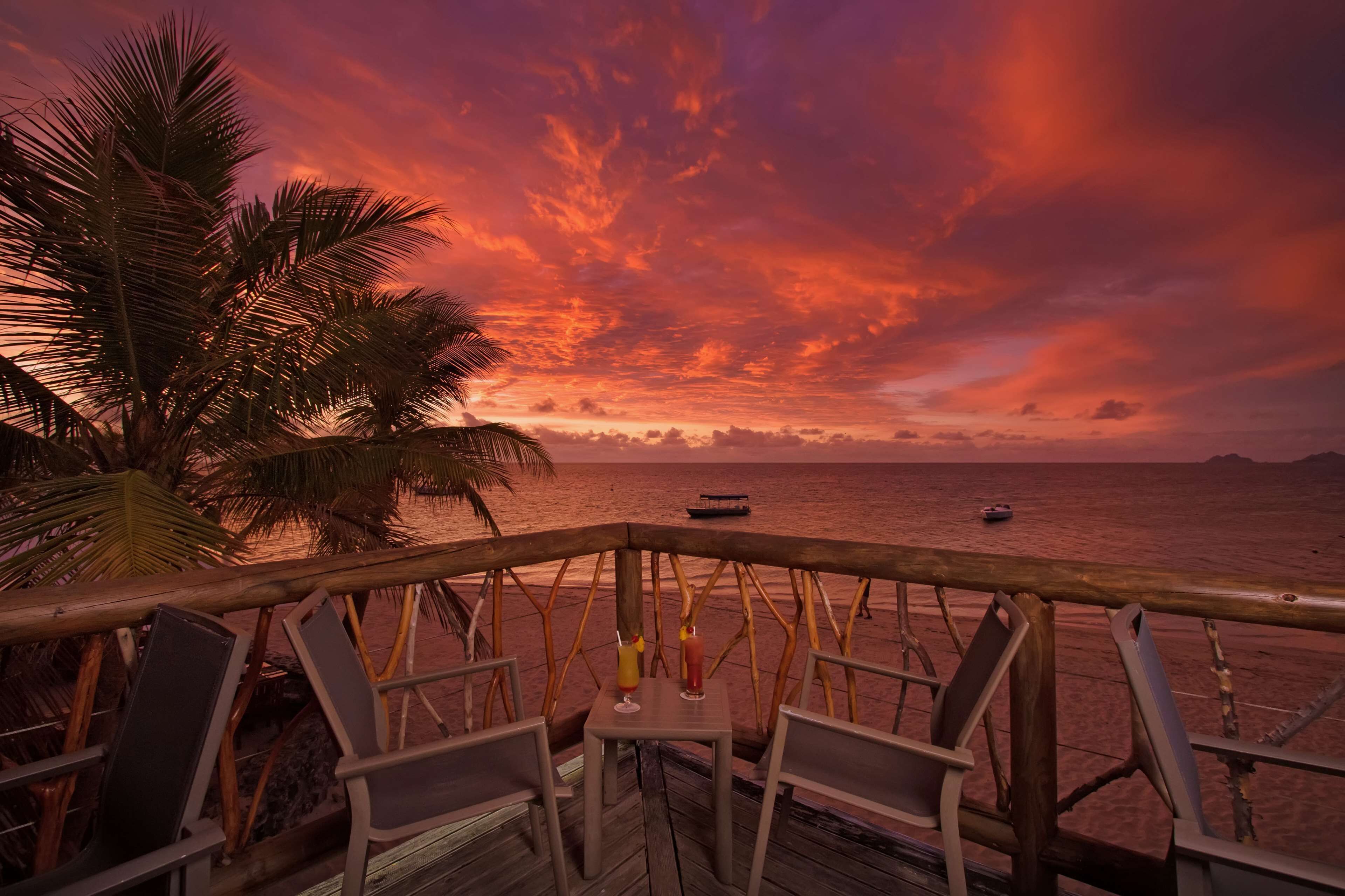 Um deck de madeira com cadeiras e uma mesa, com vista para uma praia com palmeiras e um pôr do sol.
