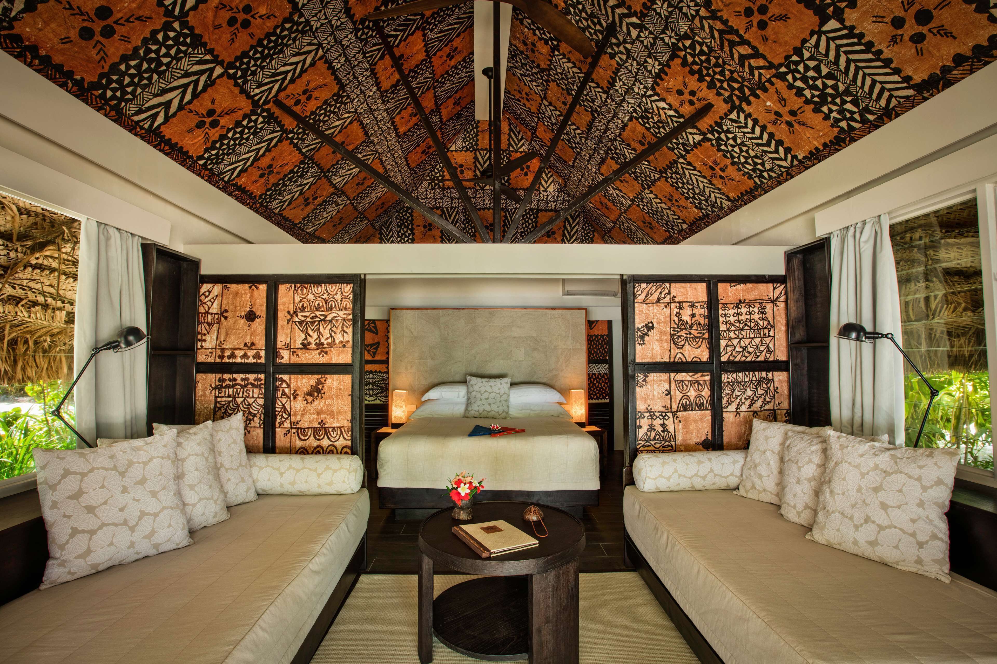 A bedroom with a large bed, two sofas, a small table, patterned ceiling, and a view of greenery.