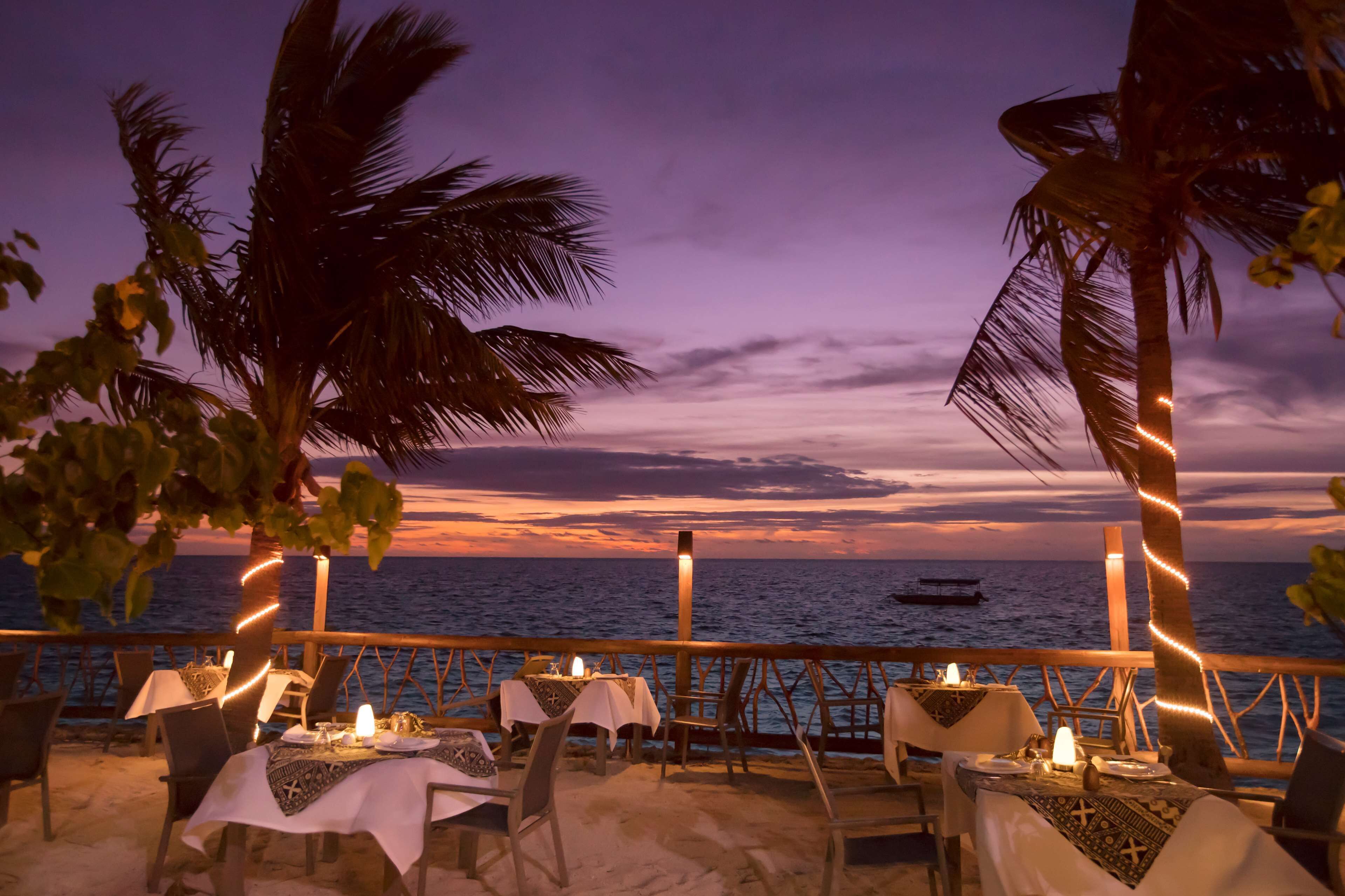 An outdoor dining area with tables set for two, overlooking the sea, with palm trees and a sunset in the background.
