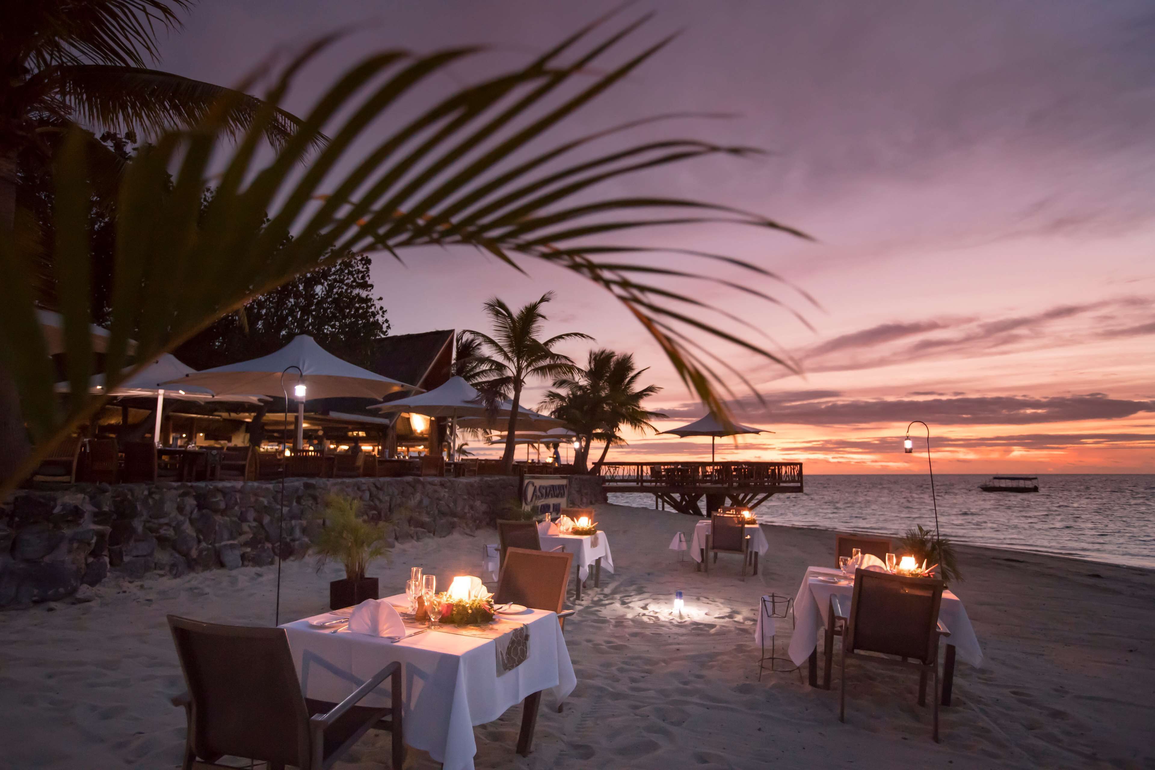 A beachfront dining area with tables set for dinner, palm trees, and a sunset view.