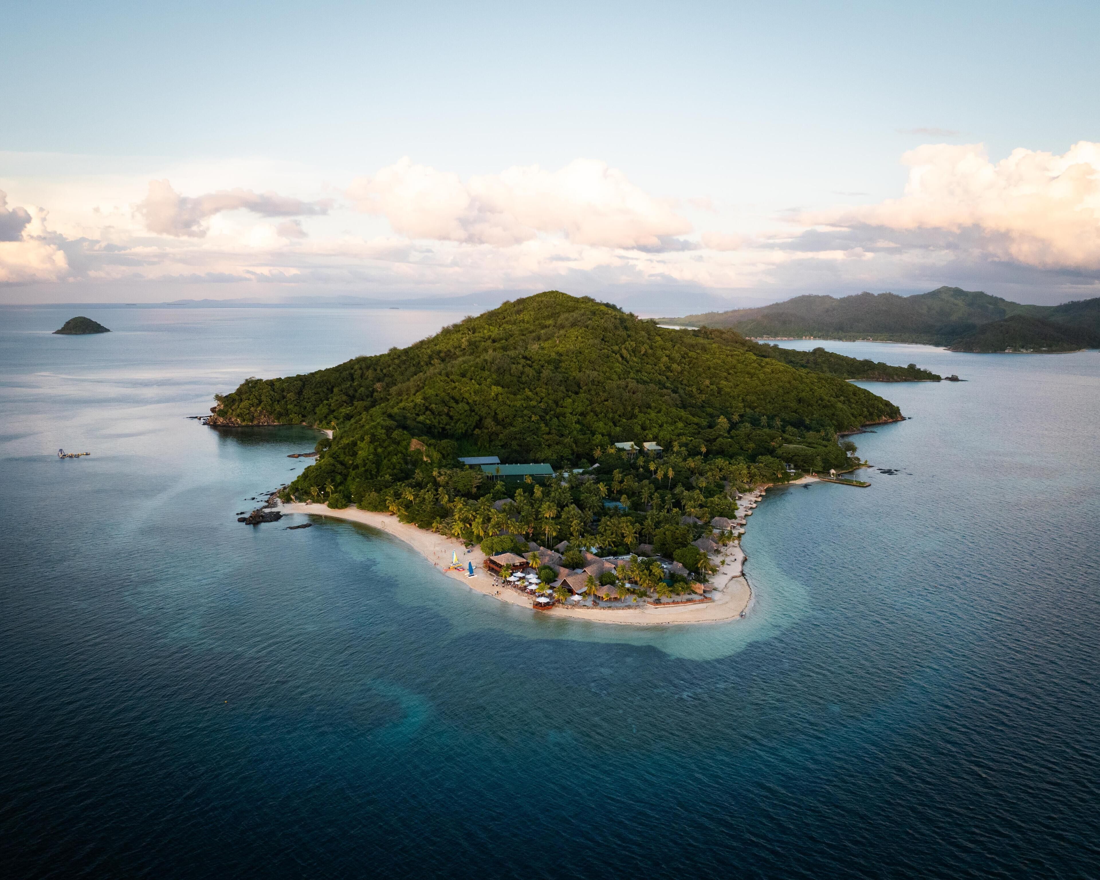 Vista aérea de una isla tropical con una pequeña zona de playa, rodeada de aguas azules cristalinas y exuberante vegetación.