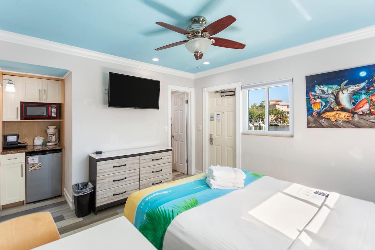 A hotel room with a bed, a desk, a microwave, a coffee maker, a TV, a ceiling fan, and a painting on the wall.