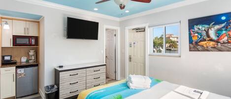 A hotel room with a bed, a desk, a microwave, a coffee maker, a TV, a ceiling fan, and a painting on the wall.