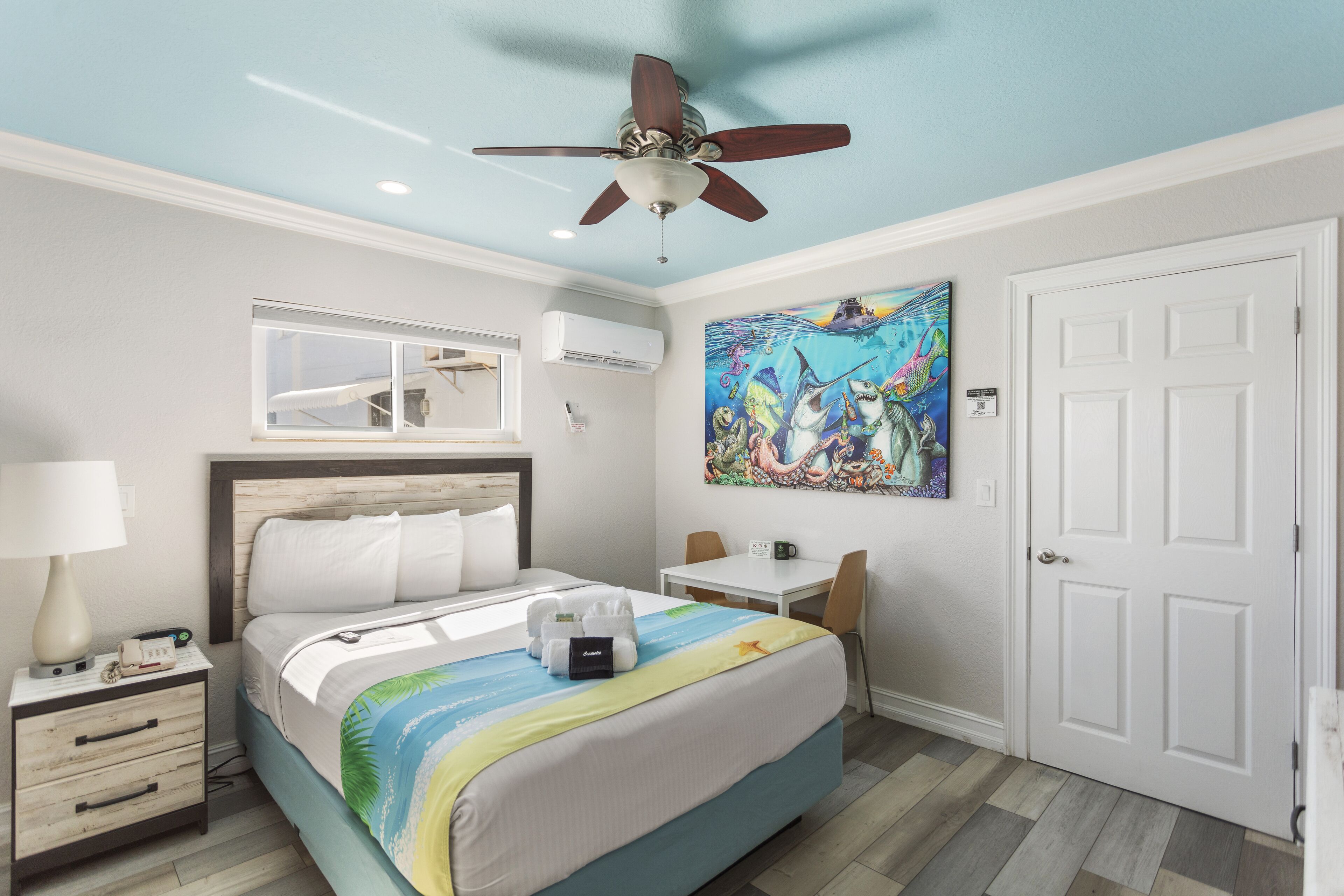 A hotel room with a bed, a desk, a chair, a ceiling fan, an air conditioning unit, and a door.