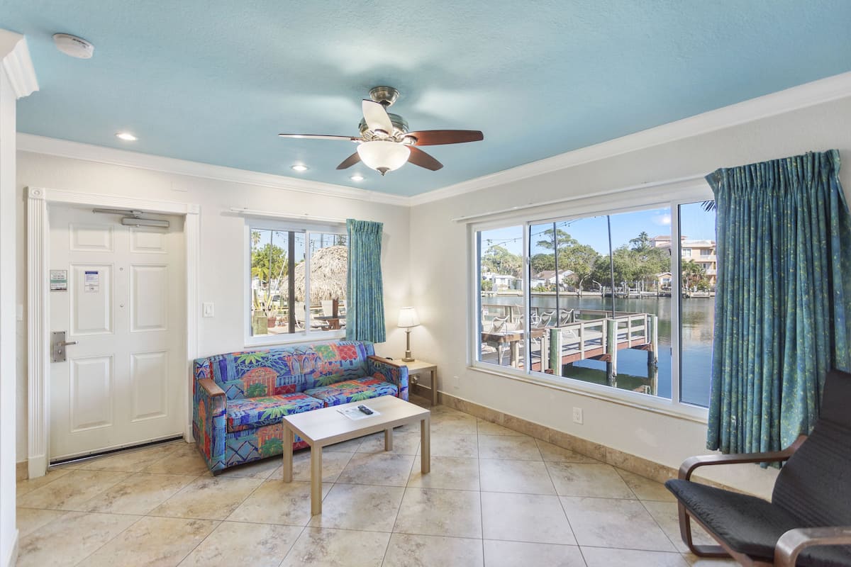 A room with a ceiling fan, a colorful sofa, a table, and large windows with curtains.