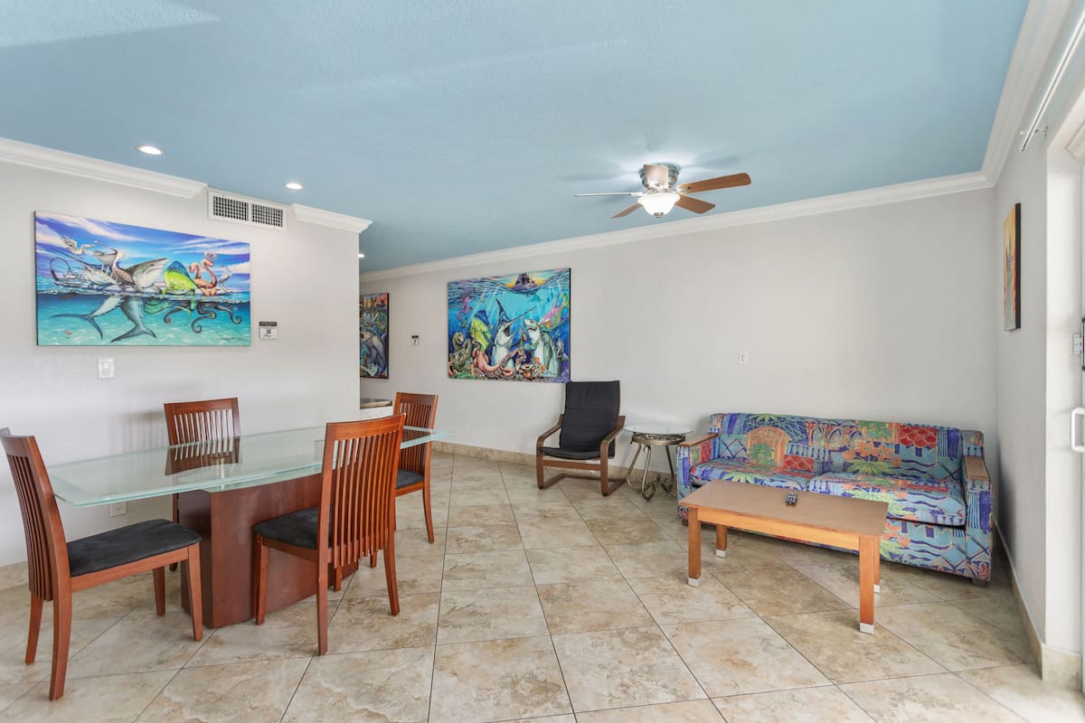 A dining area with a glass table and wooden chairs, a black armchair, a colorful sofa, and two paintings on the walls.