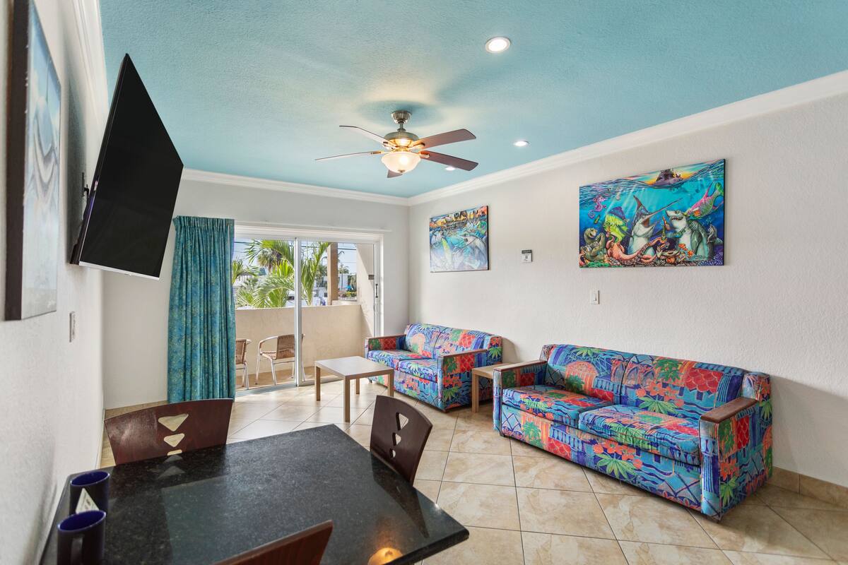 A living room with a ceiling fan, a TV mounted on the wall, two colorful sofas, a dining table with chairs, and a painting on the wall.