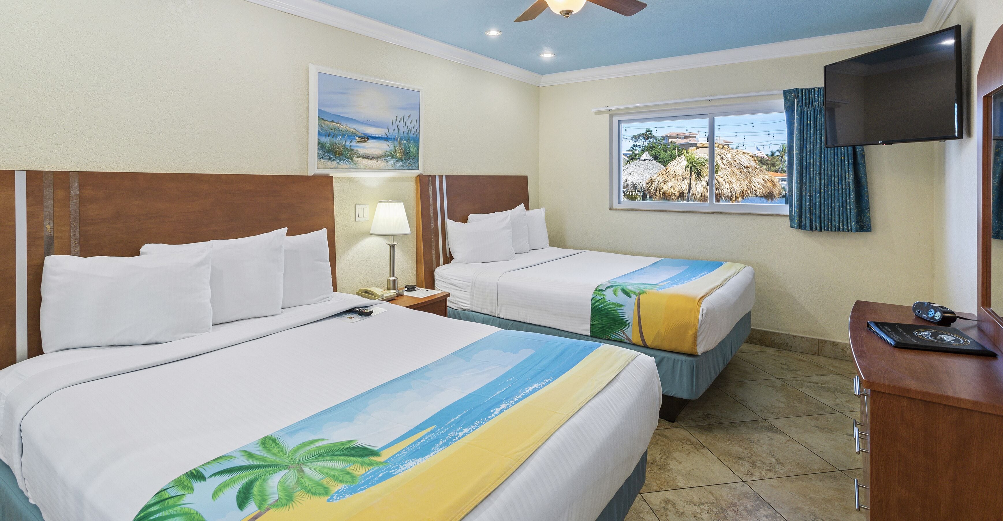Quarto de hotel com duas camas, televisão, escrivaninha e janela com vista para palmeiras e praia.