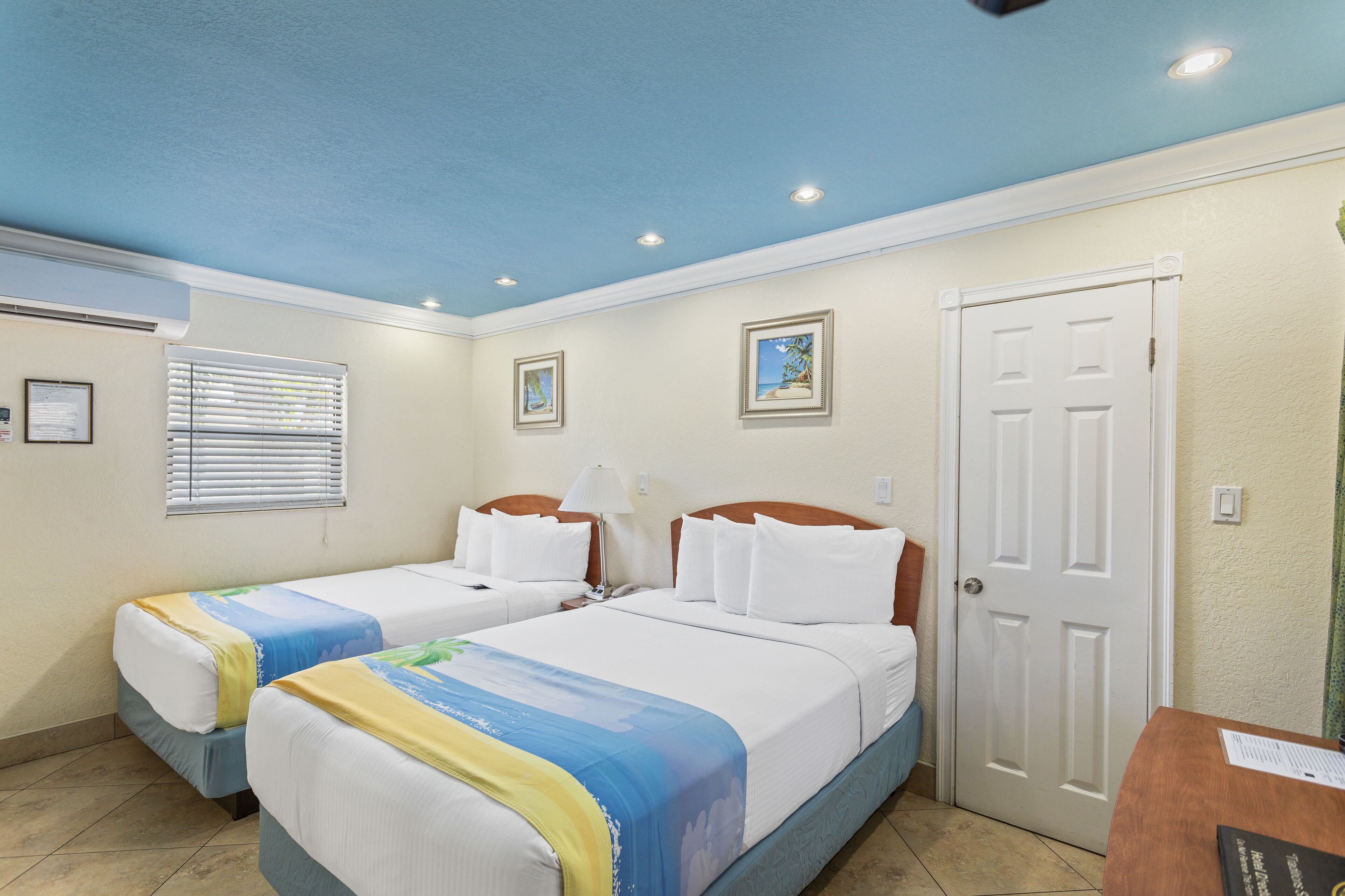A hotel room with two beds, a door, a window with blinds, and a small table.