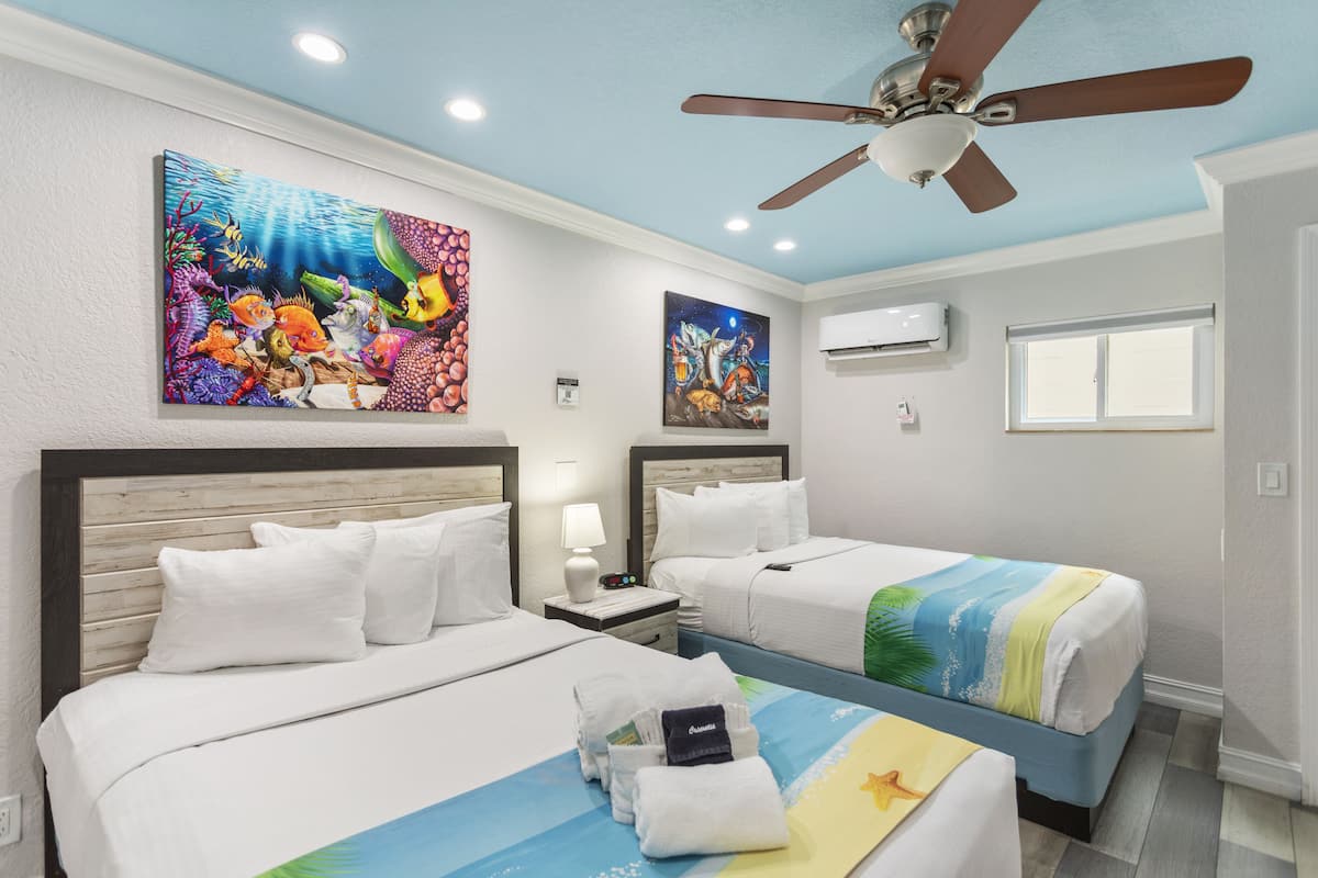 A hotel room with two beds, a ceiling fan, artwork depicting underwater scenes, and a window.