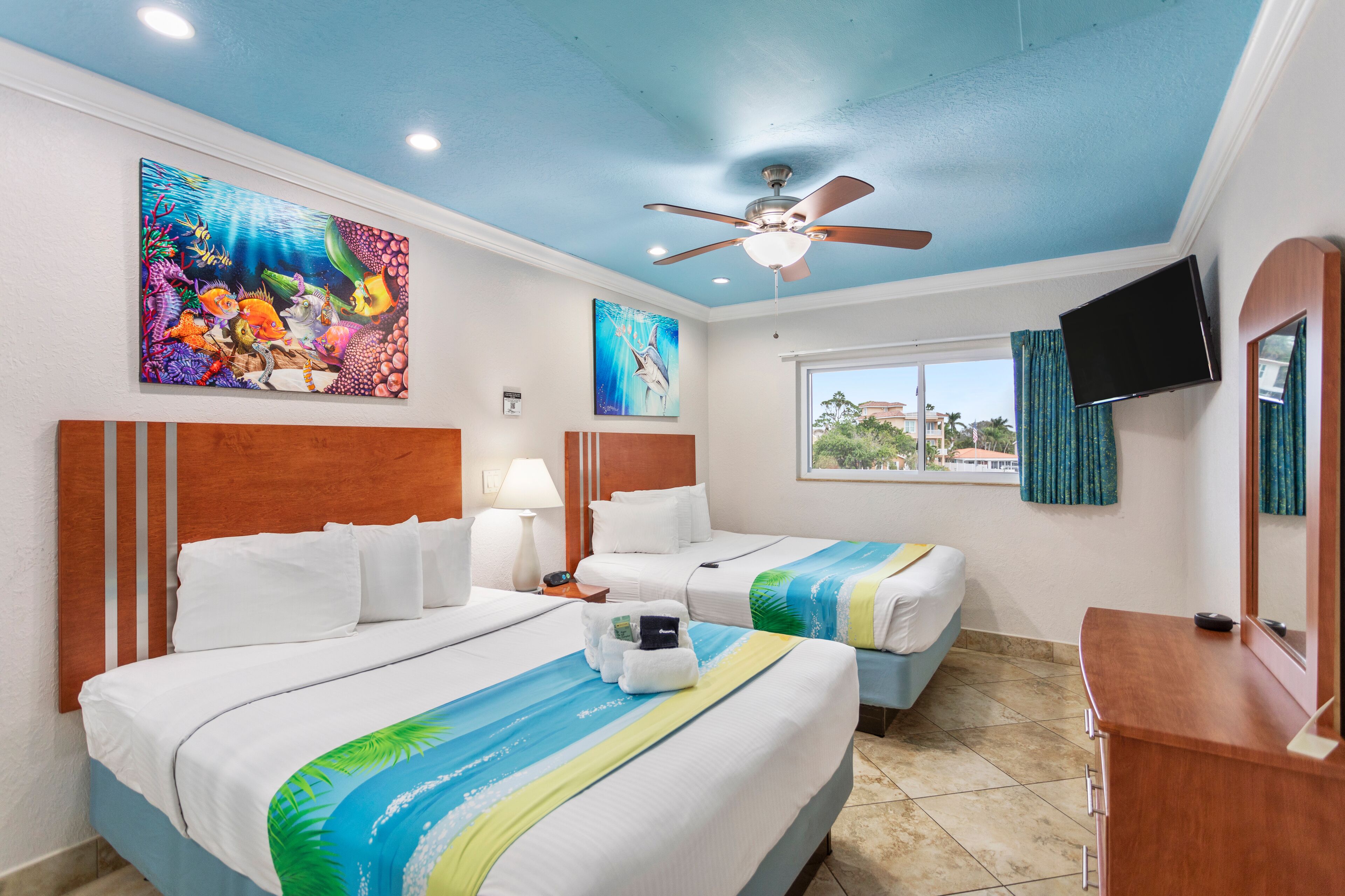 A hotel room with two beds, a ceiling fan, a TV, and artwork on the walls.