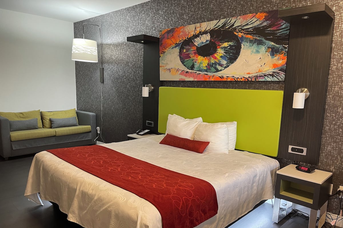 Ein Hotelzimmer mit einem großen Bett, einem grünen Kopfteil, einem grauen Sofa und einem farbenfrohen, abstrakten Gemälde über dem Bett.