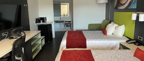 A hotel room with a desk, two beds, a TV, a kitchenette, and a painting on the wall.