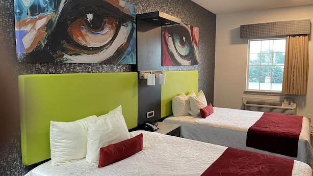 A hotel room with two beds, each with a green headboard, white bedding, and red accents. There are abstract paintings on the wall above the beds.