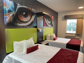 A hotel room with two beds, each with a green headboard, white bedding, and red accents. There are abstract paintings on the wall above the beds.