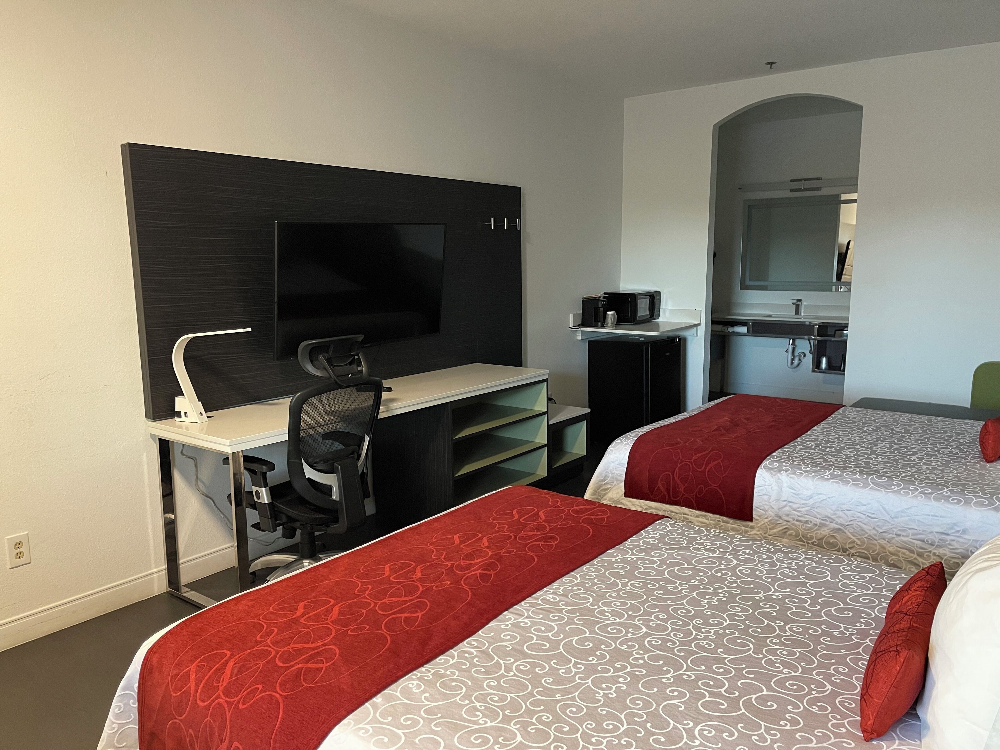 A hotel room with a desk, two beds, a TV, a microwave, and a small kitchenette.