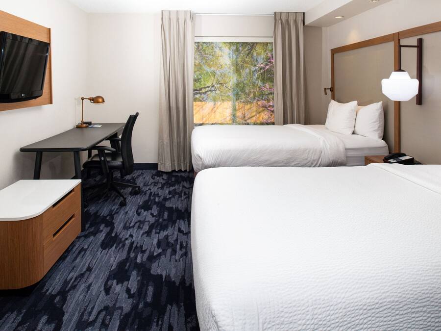 A hotel room with two beds, a desk, a chair, a TV, and a large window with curtains.
