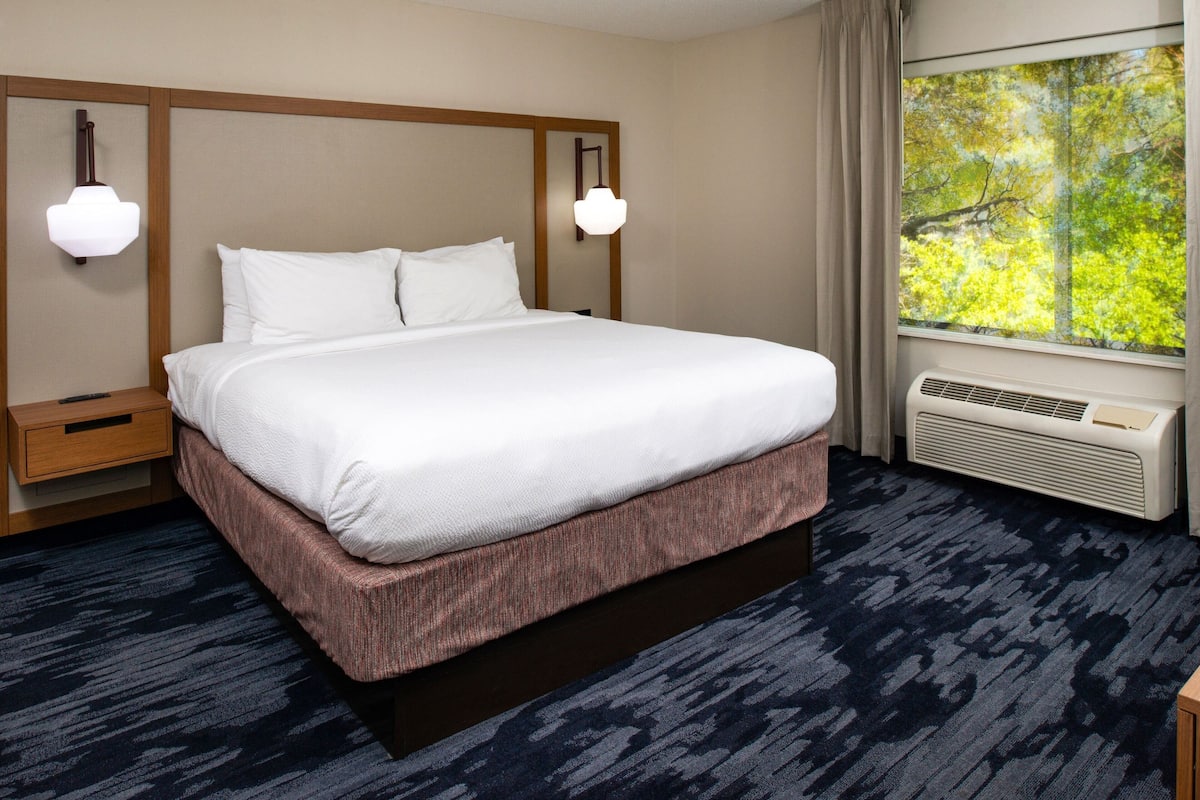 Une chambre d’hôtel avec un grand lit, deux tables de chevet, une fenêtre donnant sur des arbres et un appareil de climatisation.