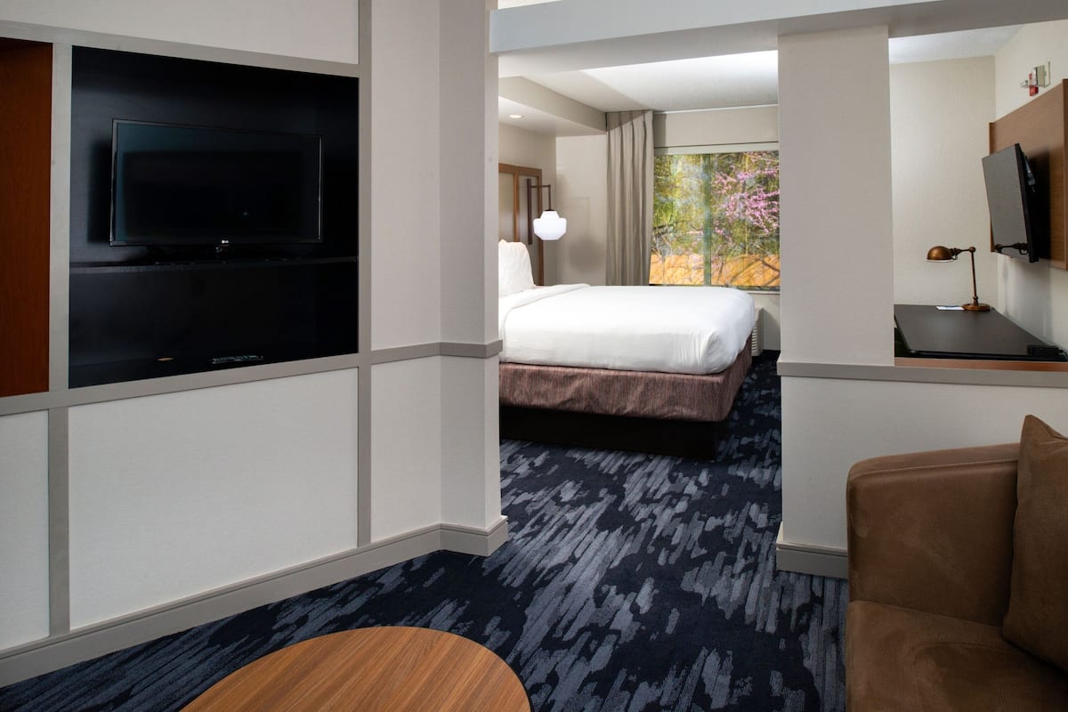 Une chambre d’hôtel dotée d’un grand téléviseur à écran plat fixé au mur, d’un lit recouvert d’une couette blanche, d’un bureau avec une lampe et offrant une vue sur les arbres par la fenêtre.