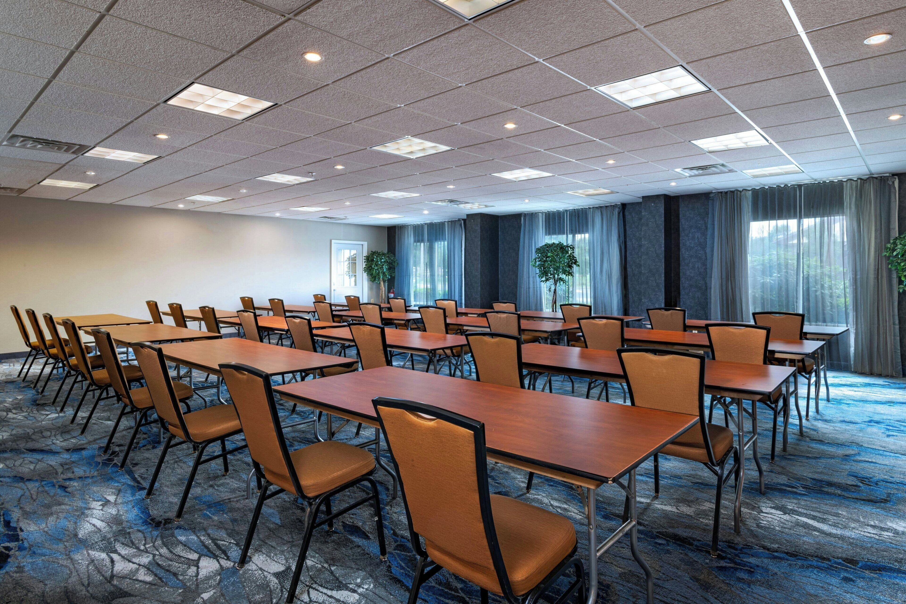 A conference room with multiple tables and chairs, large windows with curtains, and a carpeted floor.